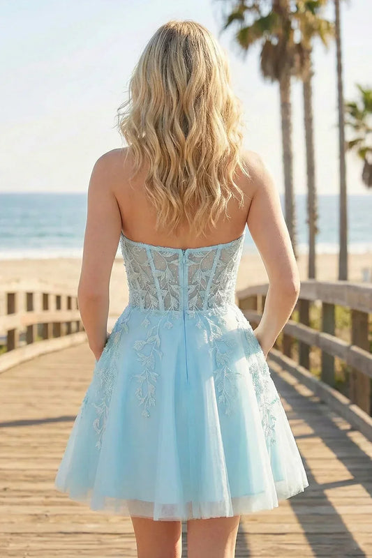 Back view of a woman in a light blue A-line tulle lace homecoming dress with floral appliqué, standing on a boardwalk by the beach.