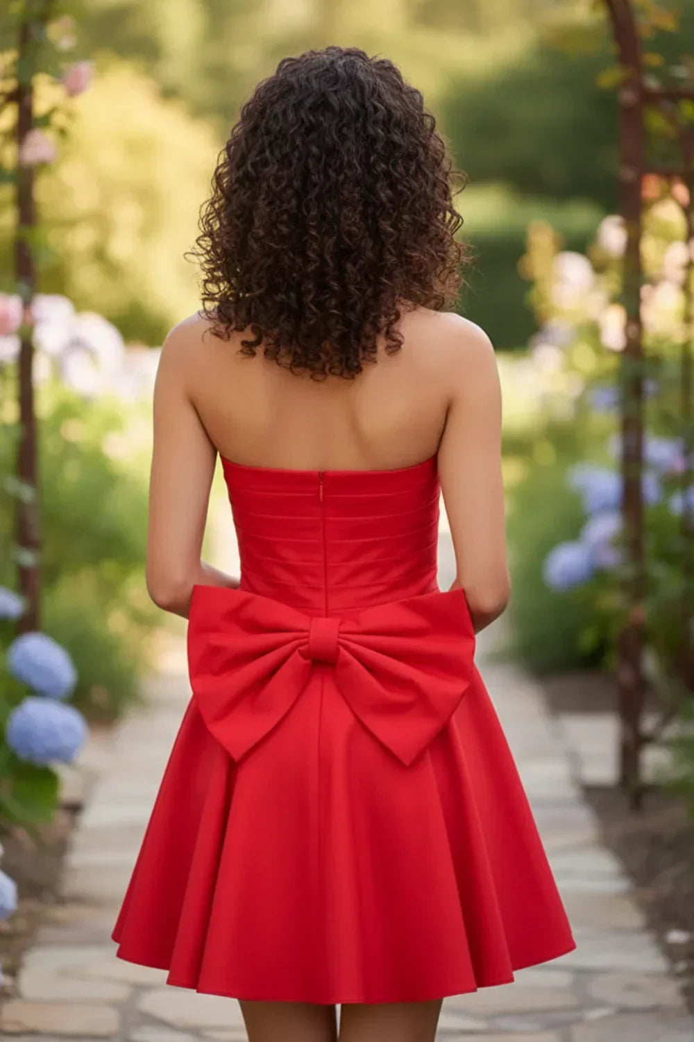 Back view of strapless red mini A-line homecoming dress with a large bow, outdoor garden