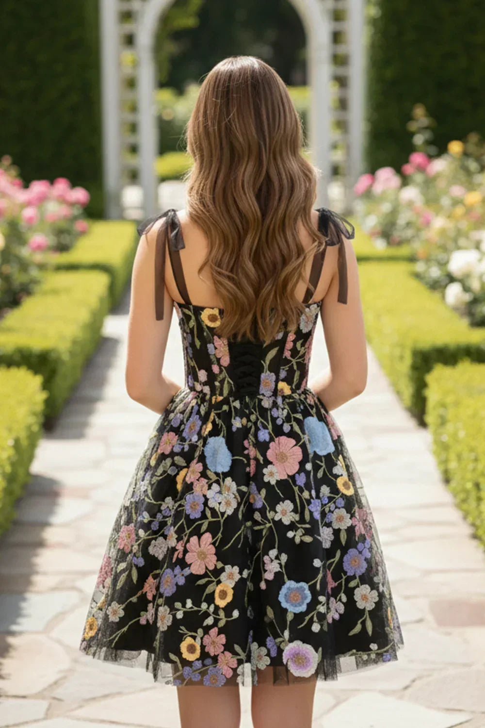Back view of a woman in a black A-line tulle short homecoming dress with colorful floral embroidery and 3D flowers, standing in a garden pathway.