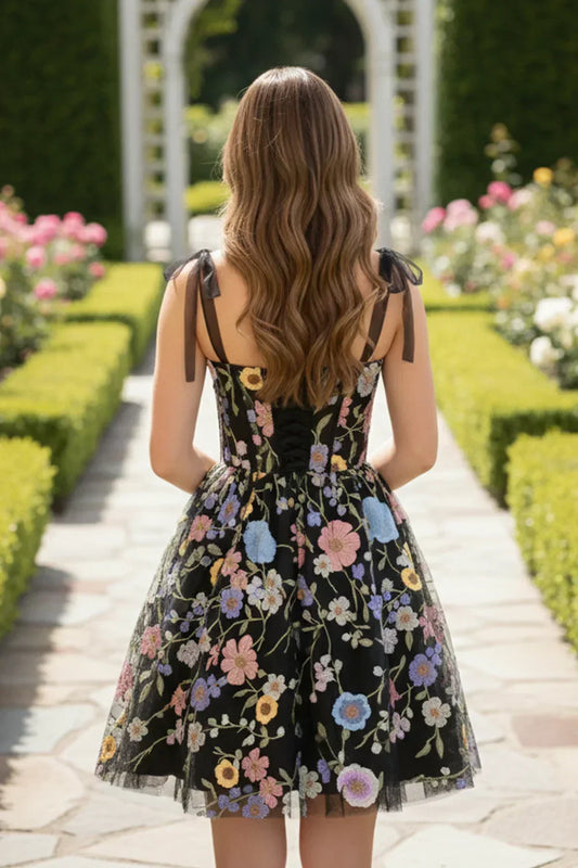 Back view of a woman in a black A-line tulle short homecoming dress with colorful floral embroidery and 3D flowers, standing in a garden pathway.
