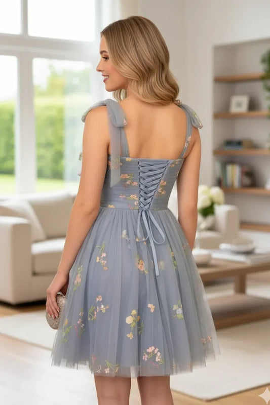 Back view of a woman in a blue A-line tulle short homecoming dress with floral embroidery and lace-up detail, standing in a modern living room.