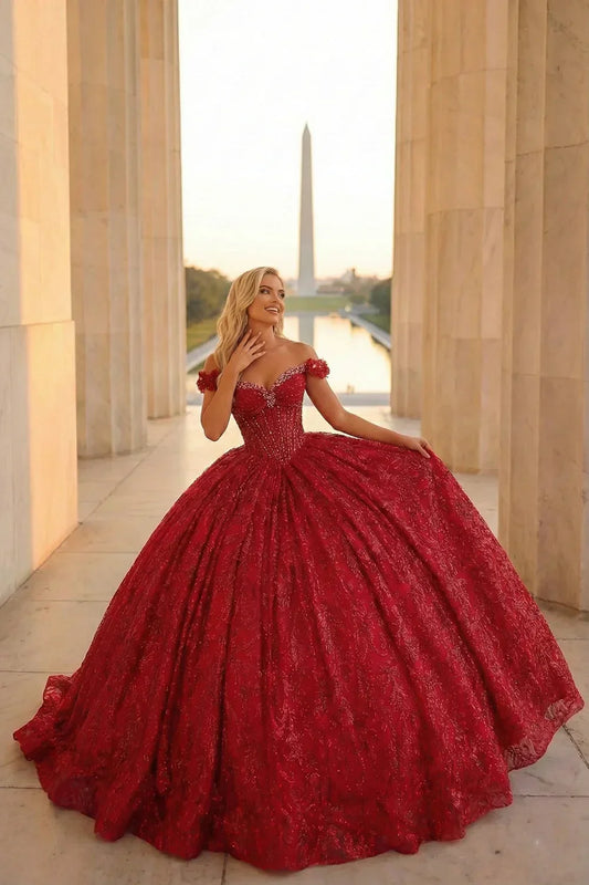Woman in elegant red ball gown with off-shoulder detail, standing outdoors at sunset
