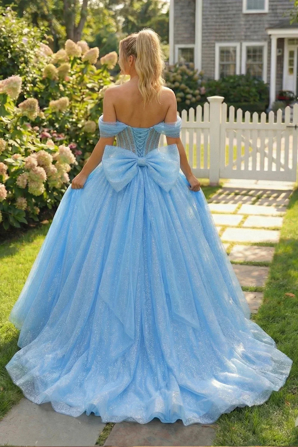 Woman in sparkling blue ball gown with off-shoulder sleeves and large bow, standing in a garden