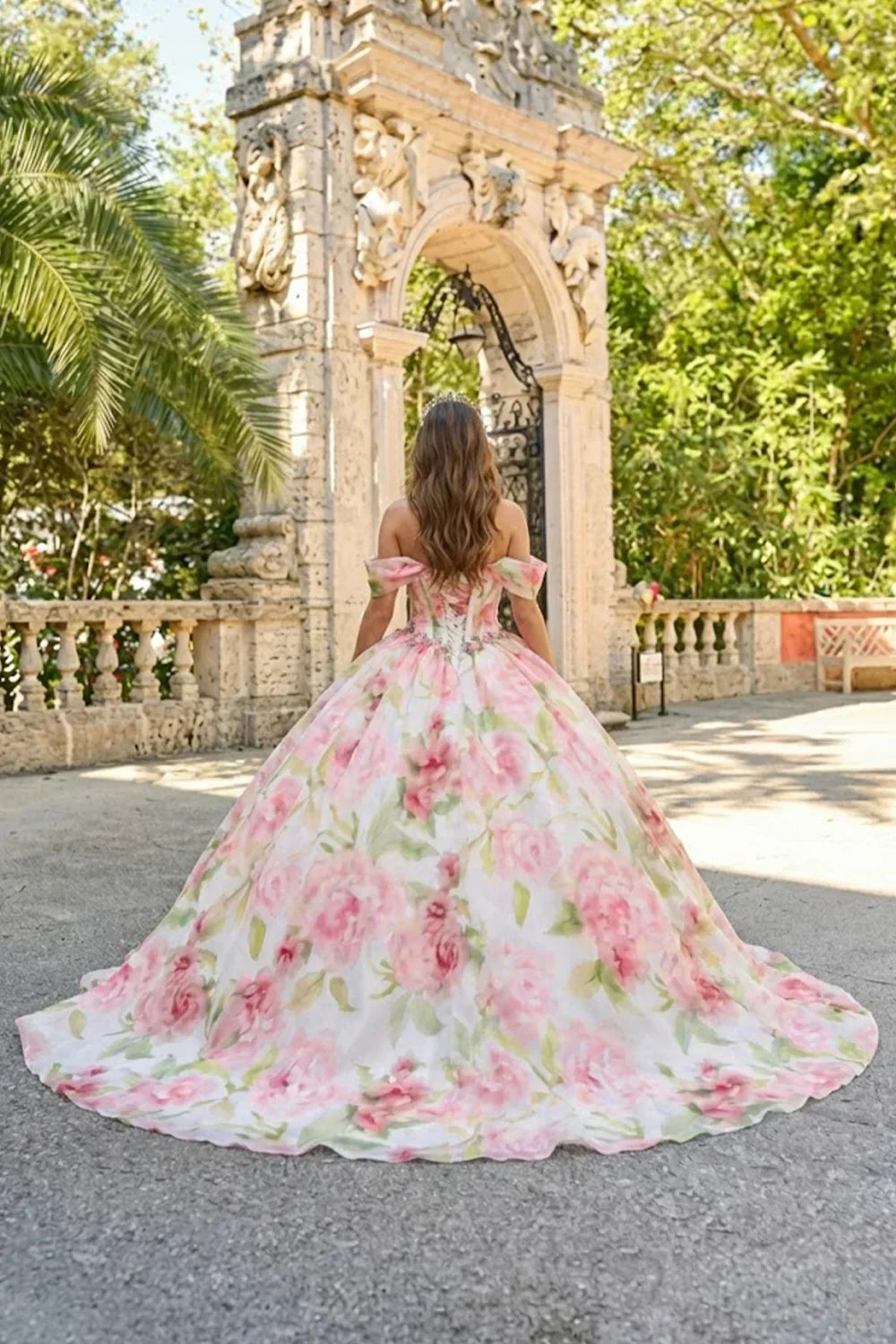 Elegant ball gown with pink floral pattern and cold-shoulder design, worn outdoors by an archway