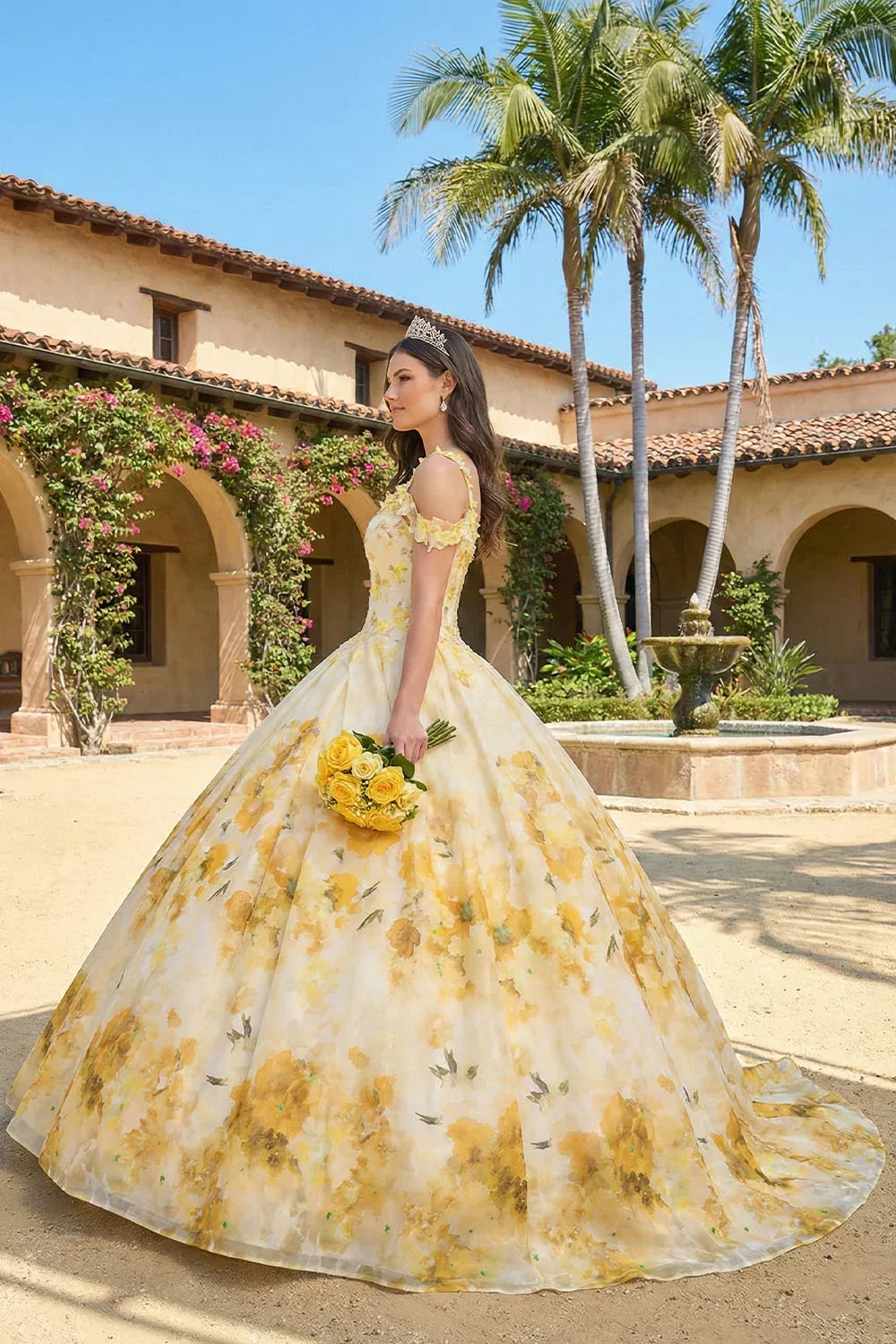 Woman in floral yellow ball gown holding yellow roses, wearing tiara, outdoors in courtyard
