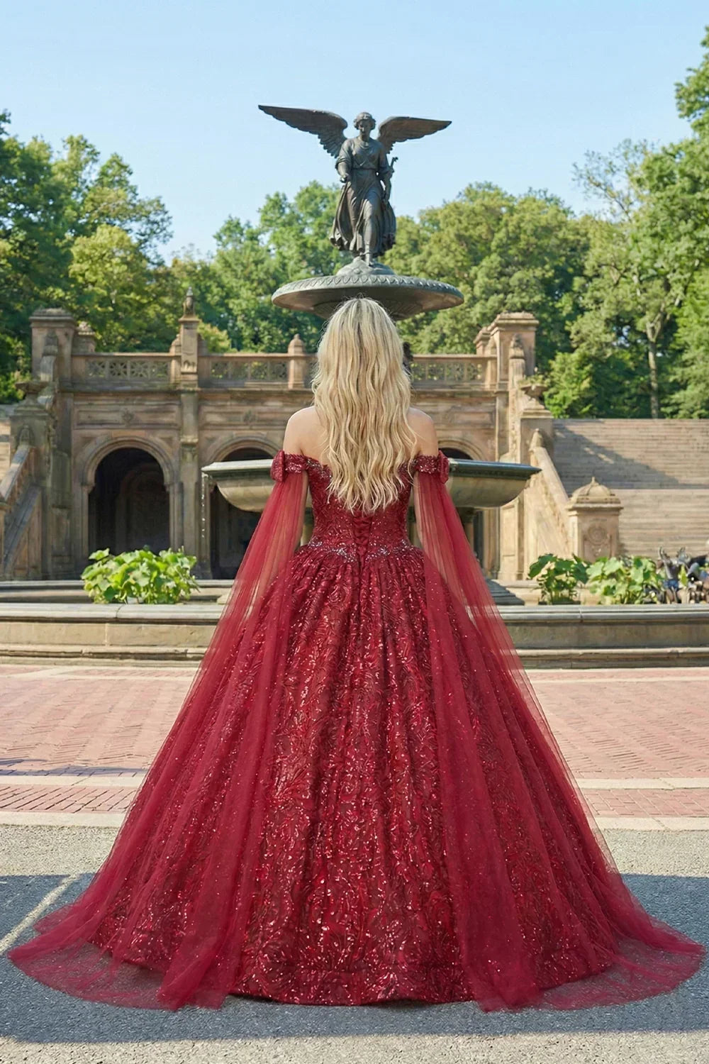 Woman in glittery red ball gown with sheer cape, standing outdoors by a stone fountain