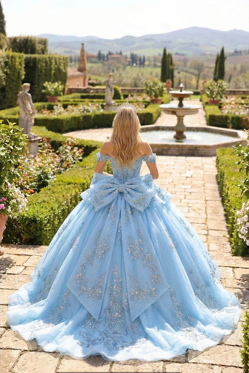 Layered ruffled blue ball gown with embroidery, cold shoulders, and large bow in garden setting