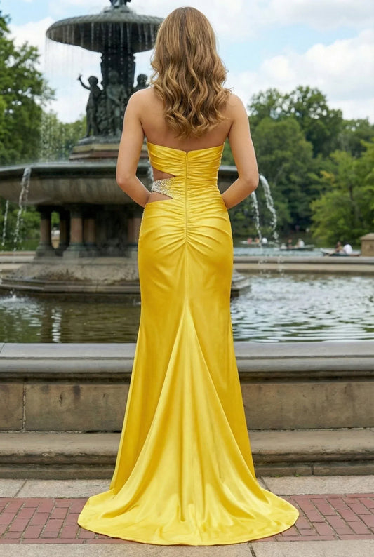 Woman in a strapless yellow prom dress with cutout and ruched back, standing by a fountain