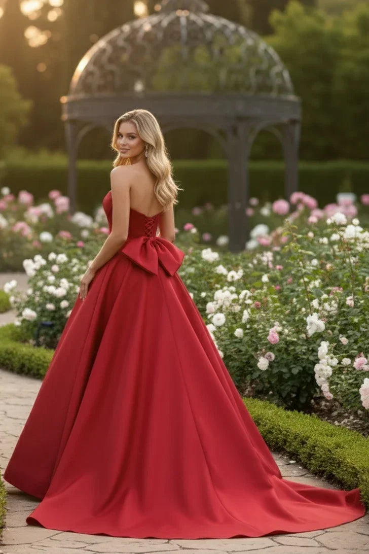 Woman in a red A-line satin prom dress with sweetheart neckline and large bow back in a garden.