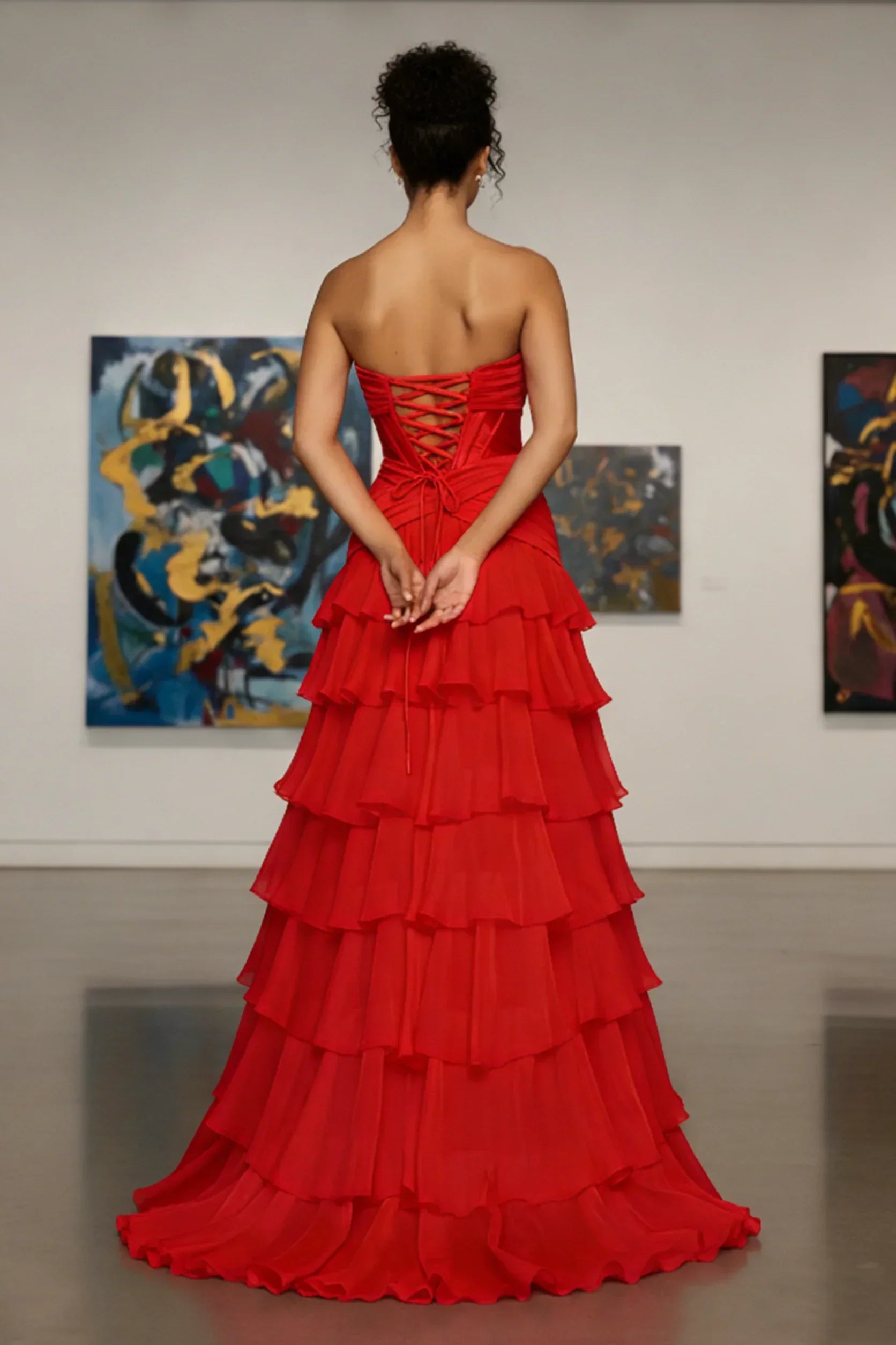 Woman in a strapless red tiered evening gown with lace-up back, standing in an art gallery.