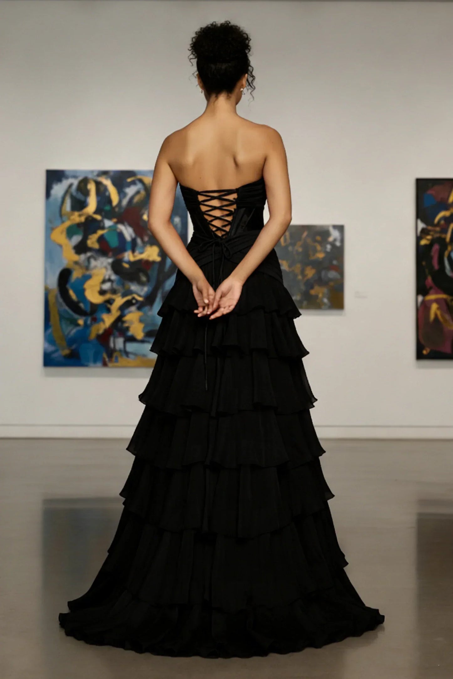 Back view of woman in strapless black tiered evening gown with lace-up corset bodice, standing in art gallery