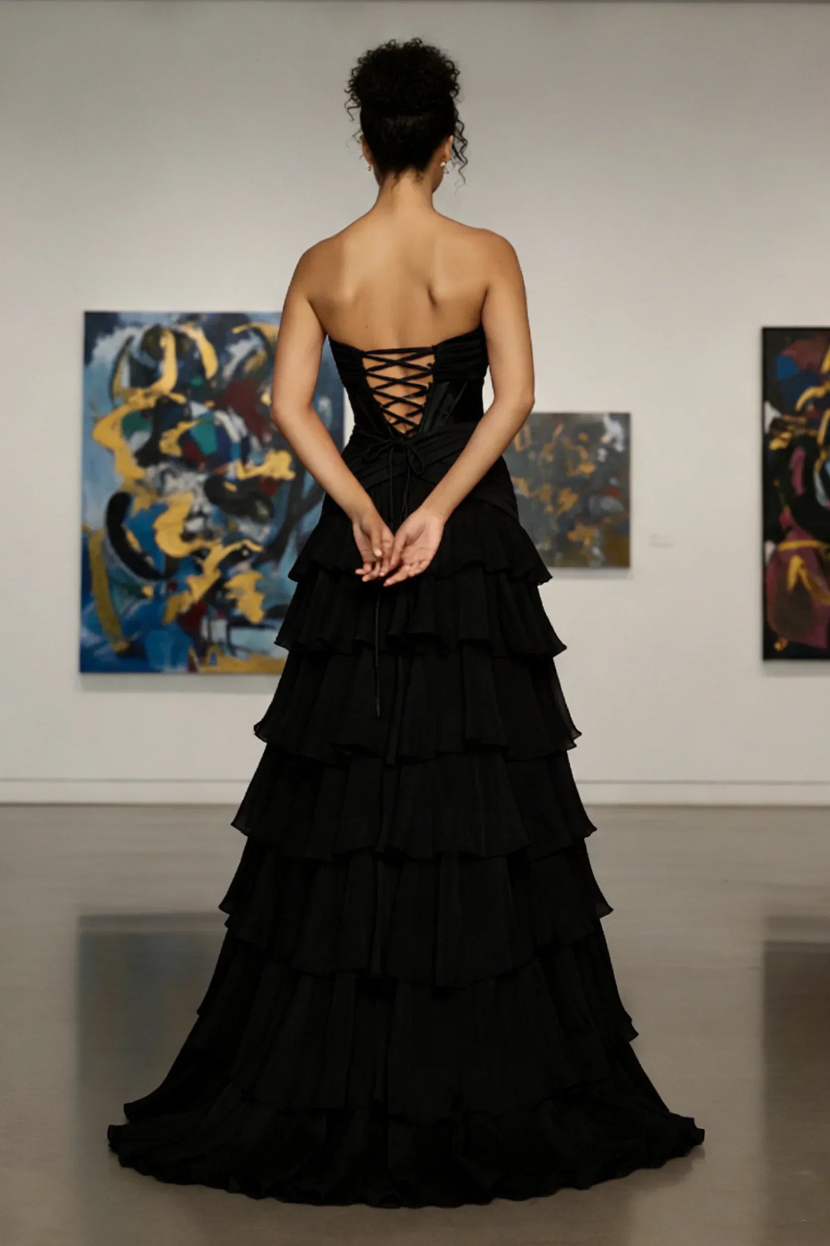 Back view of woman in strapless black tiered evening gown with lace-up corset bodice, standing in art gallery