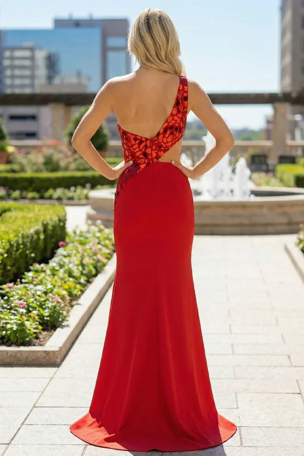 Back view of Sevoba red evening gown showing asymmetric mosaic sequin back design and side cutout, outdoor background