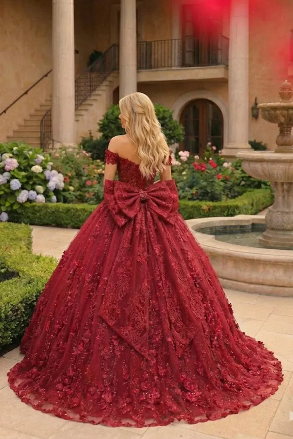 Woman in an elegant burgundy ball gown with lace, floral details, and a large bow, standing in a garden.