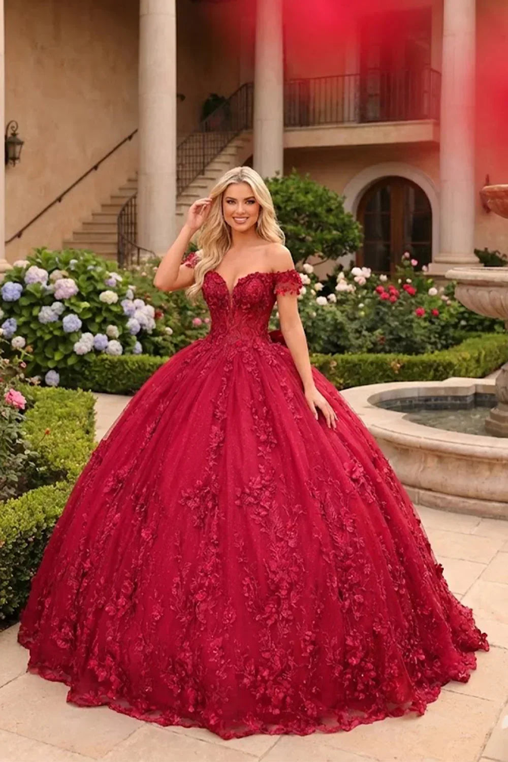 Woman in elegant red ball gown with floral lace and off-shoulder design in garden setting