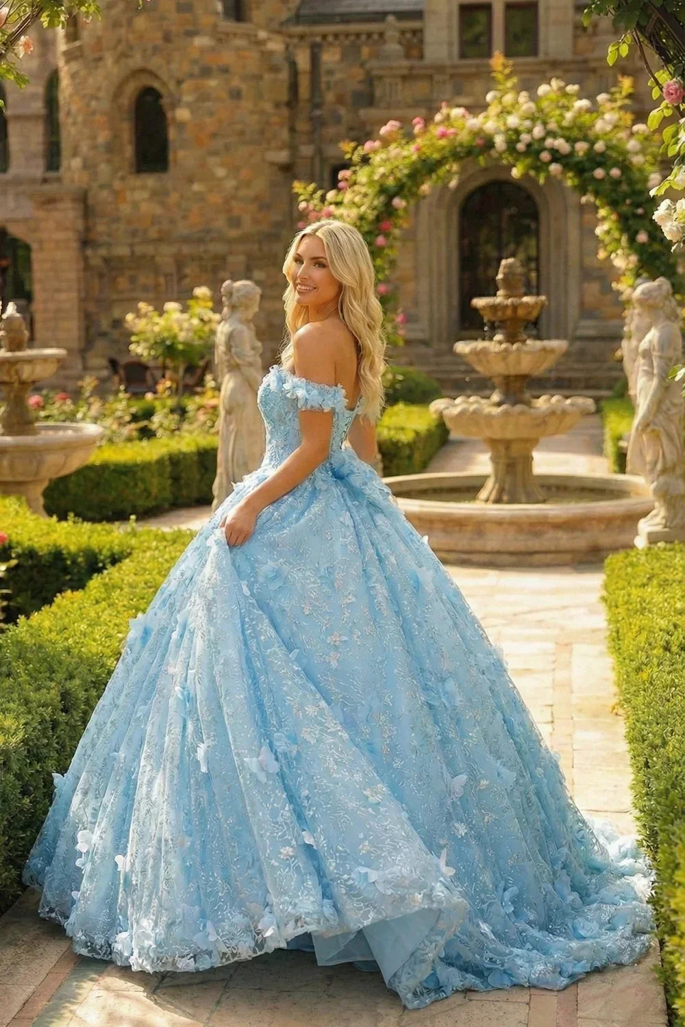 Woman in a blue ball gown with 3D floral details and off-shoulder sleeves, posing outdoors by a fountain and garden archway.