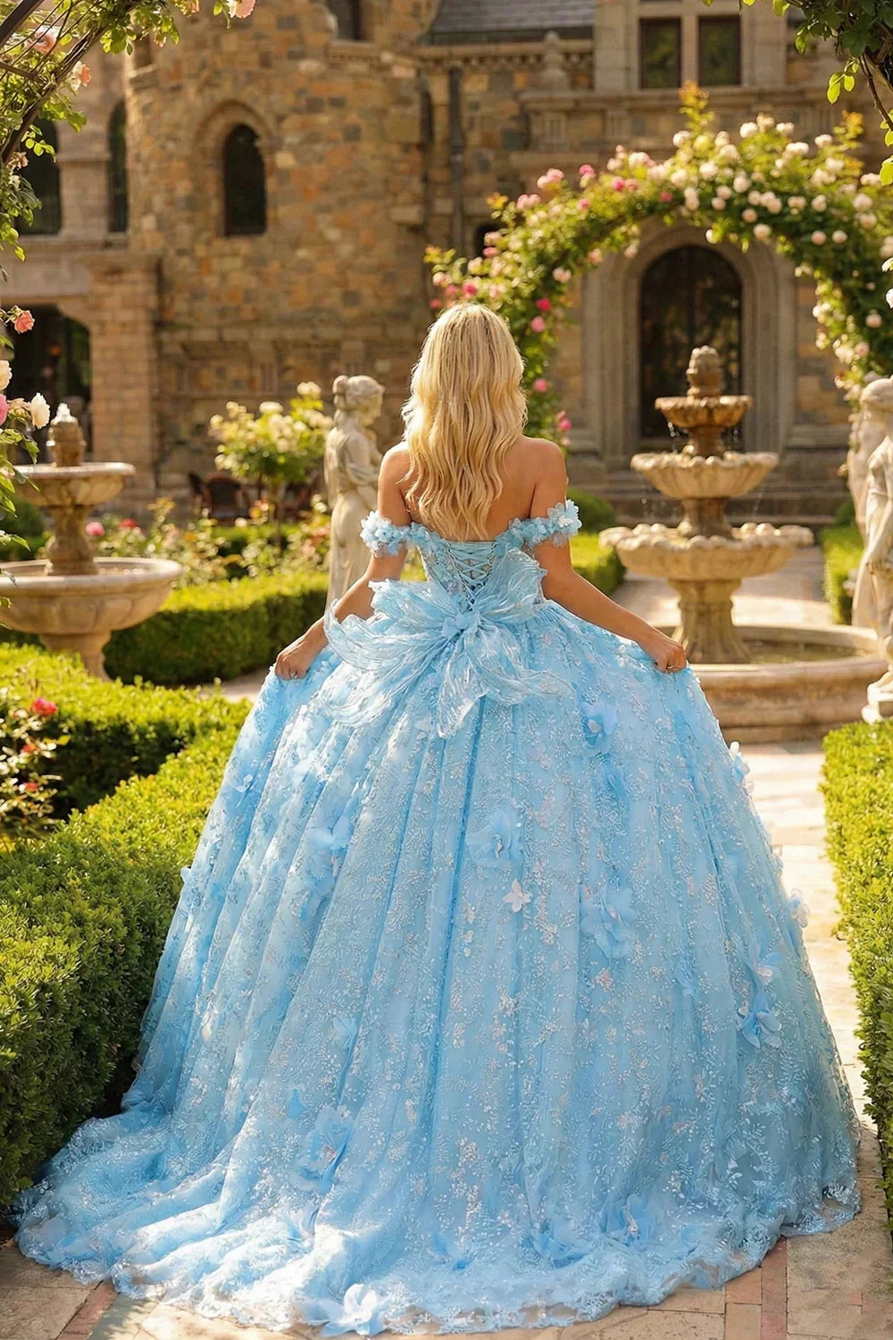 Woman in blue ball gown with 3D floral details and bow, standing in a garden.