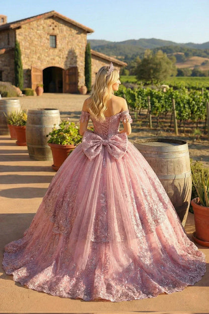 Woman in a pink cold-shoulder ball gown with floral appliqués and a large bow, outdoors near a stone villa.