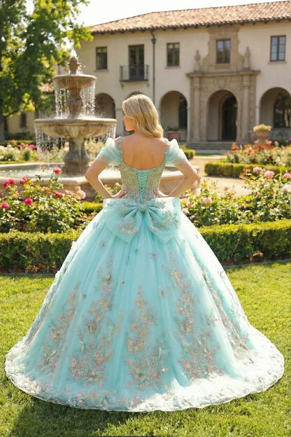 Elegant ball gown quinceañera dress with cold shoulder sleeves, floral embroidery, and a large bow, worn outdoors near a fountain.