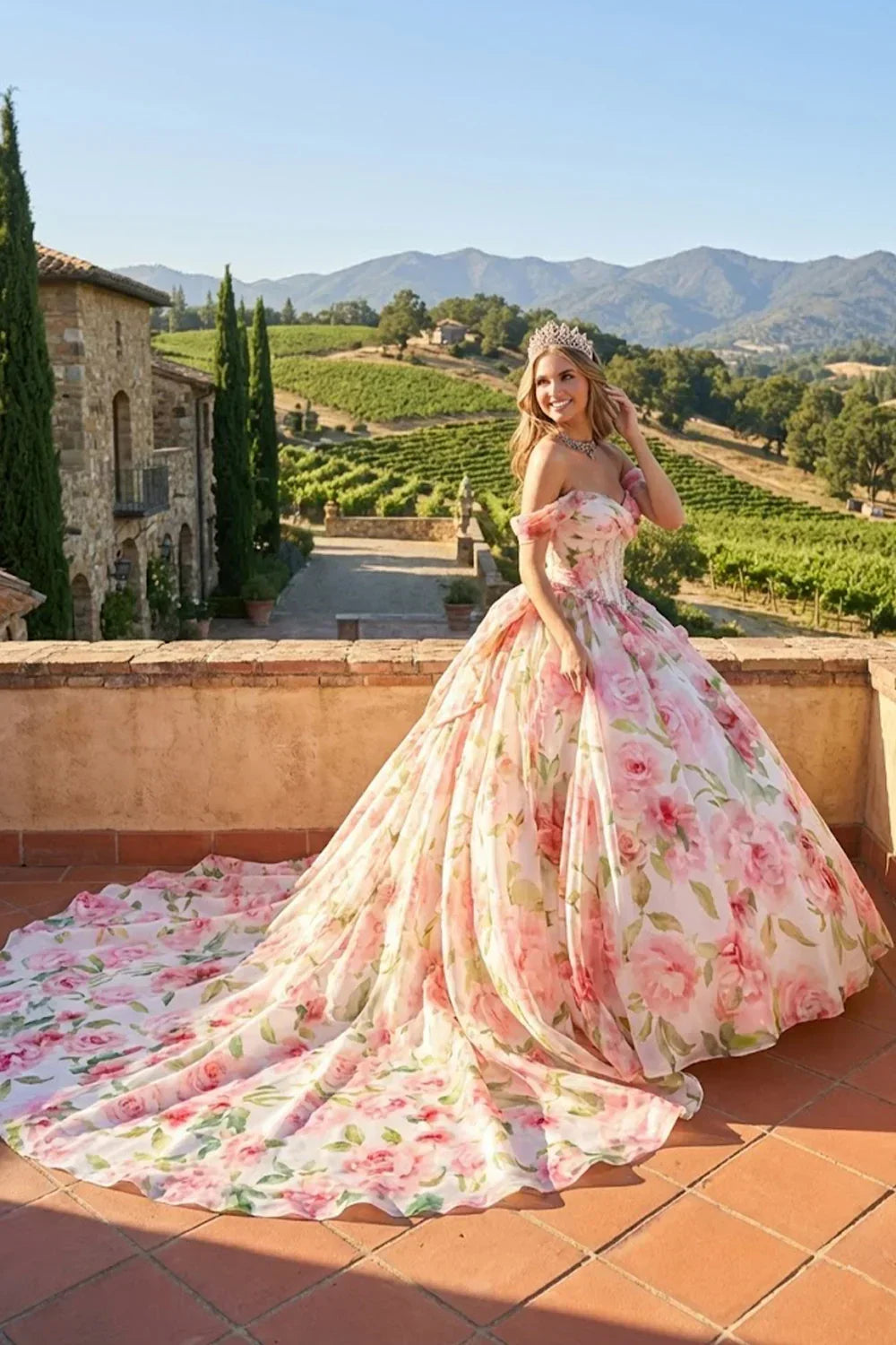 Woman in an elegant floral ball gown with cold shoulders and tiara on a scenic terrace.