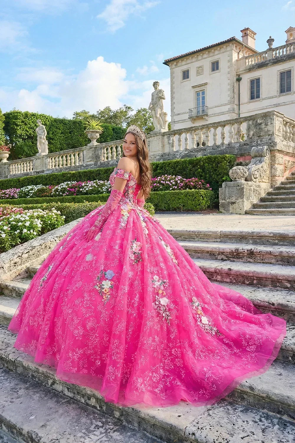 Woman in pink floral ball gown with cold-shoulder sleeves and tiara on stone steps outdoors