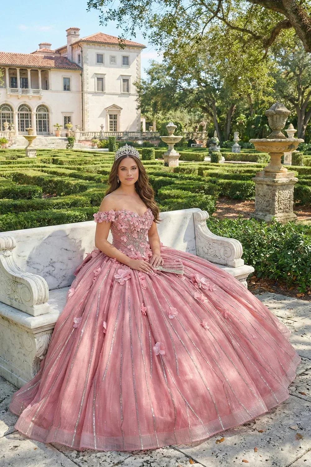 Young woman in pink off-the-shoulder ball gown with 3D floral lace, tiara, in formal garden