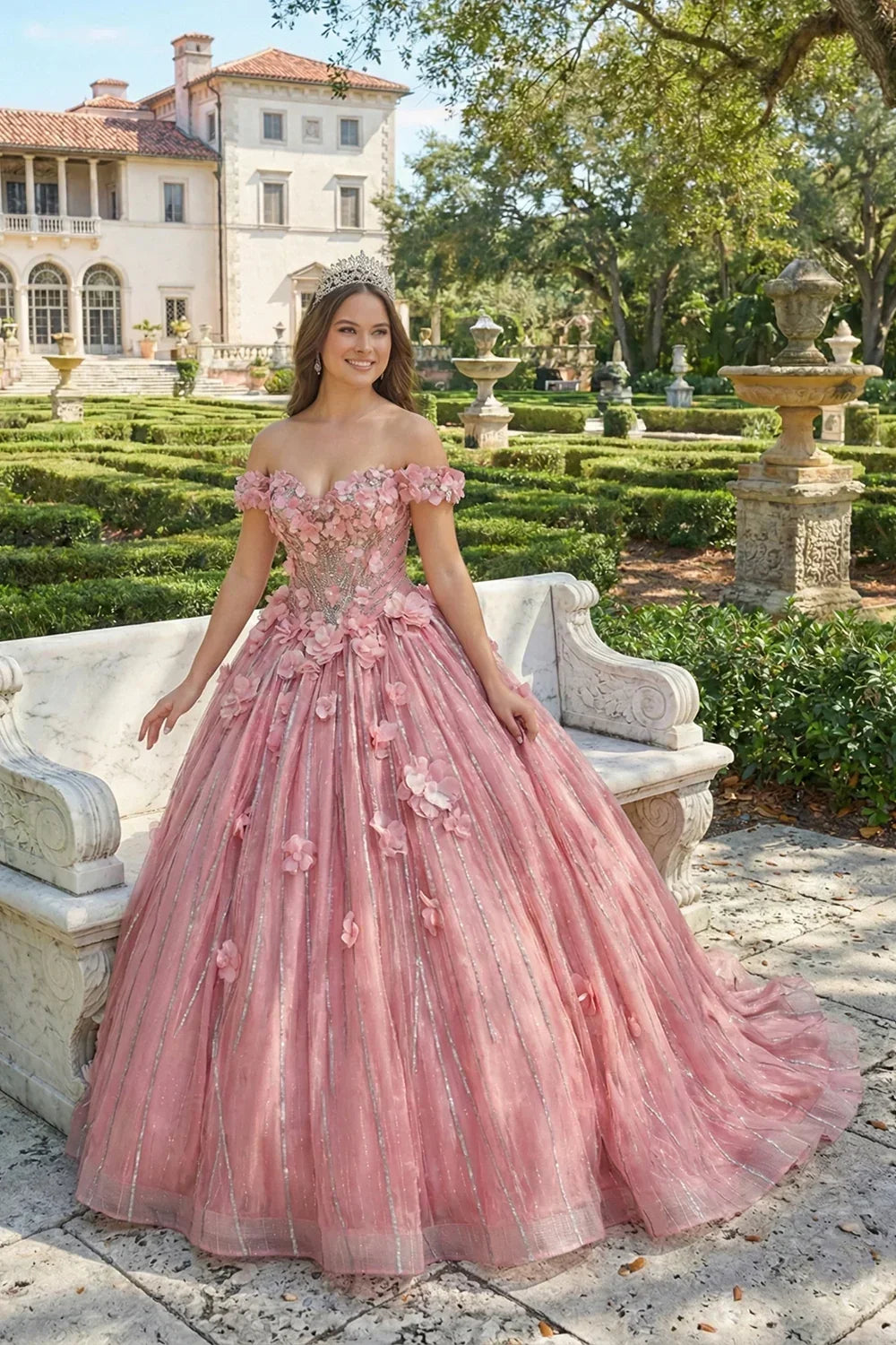 Woman in pink off-the-shoulder ball gown with 3D floral details and tiara in garden setting