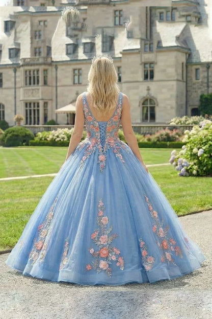 Woman in an elegant blue ball gown with floral lace, standing in a garden by a mansion