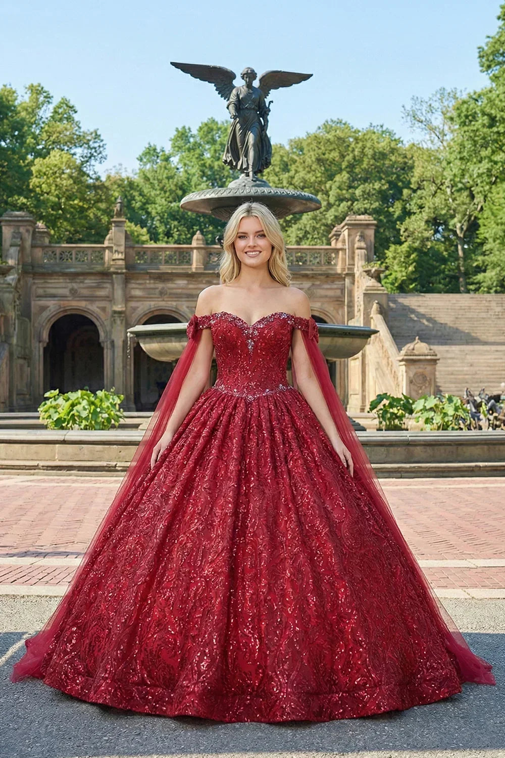 Woman in a glittery red ball gown with cold shoulders and sheer cape, posing outdoors.