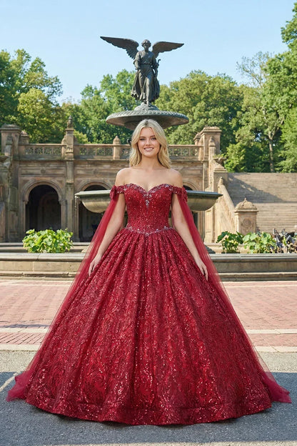 Woman in a glittery red ball gown with cold shoulders and sheer cape, posing outdoors.