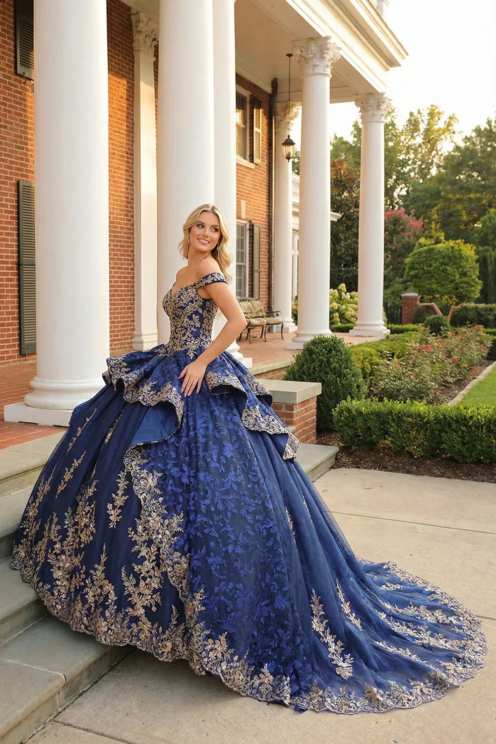 Woman in a navy blue layered ball gown with gold floral embroidery and off-shoulder straps outside a mansion.