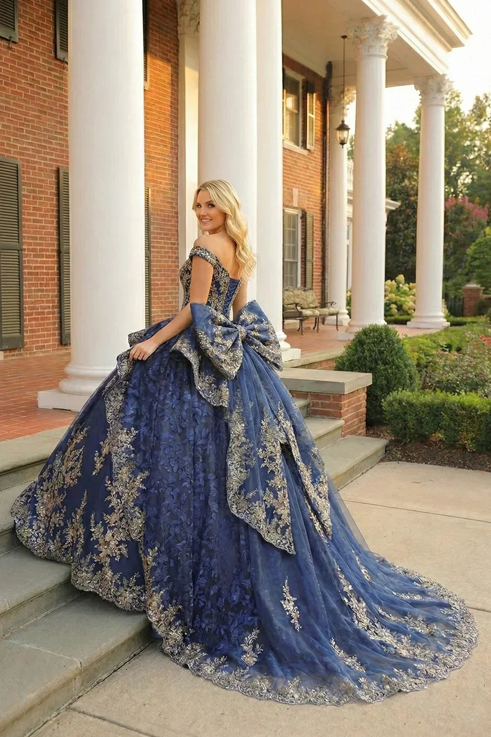 Blue layered ruffled ball gown with gold floral embroidery, off-shoulder design, and large bow, worn outdoors.