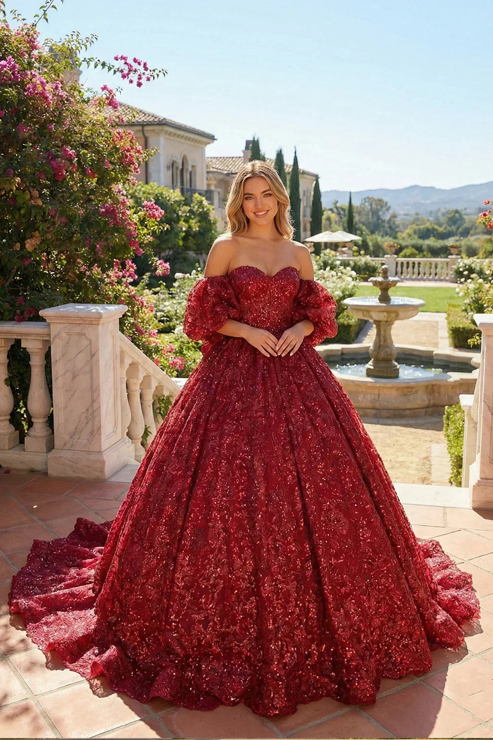 Woman in a red embroidered ball gown with off-shoulder sleeves, standing outdoors by a fountain.