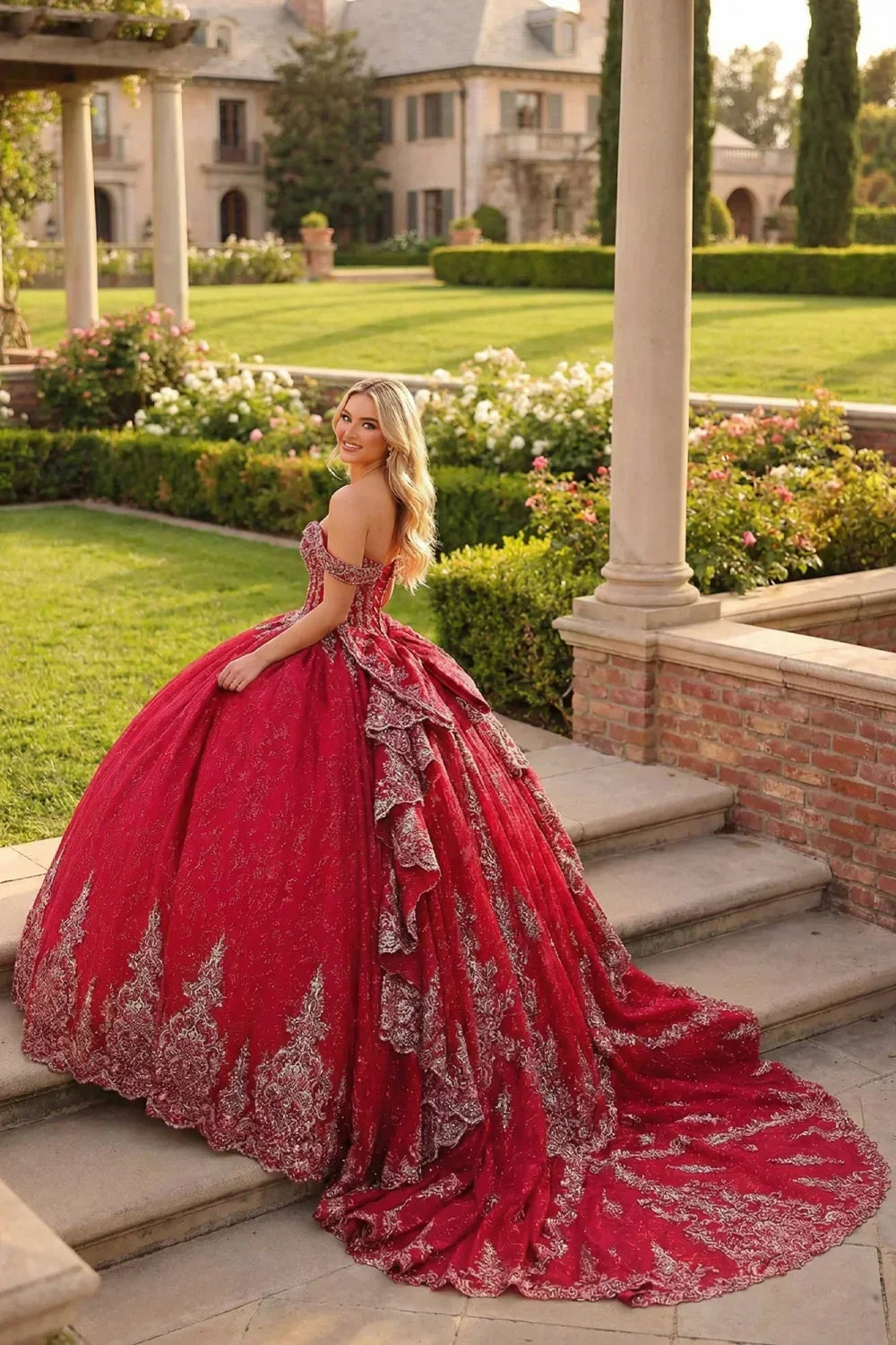 Woman in a red embroidered ball gown with train and bow, posing in a formal garden setting.