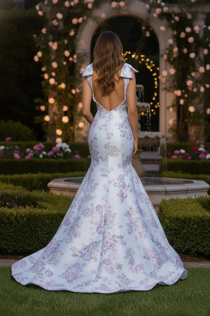 Back view of a woman in a long floral mermaid dress with bow straps in a garden setting.