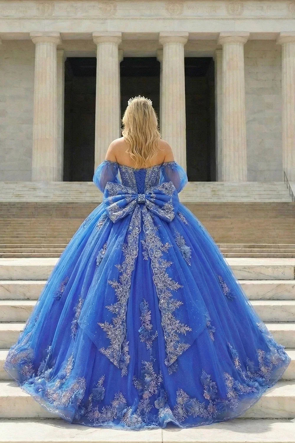 Woman in blue off-shoulder ball gown with silver floral embroidery and large bow on stairs