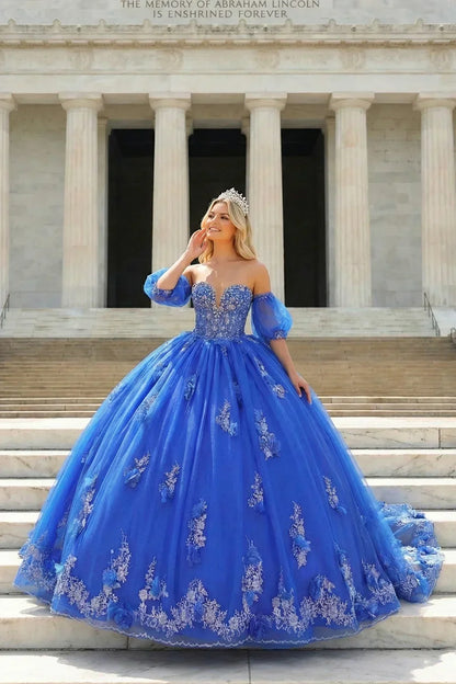 Woman in royal blue off-shoulder ball gown with silver floral embroidery and tiara, standing on marble steps.