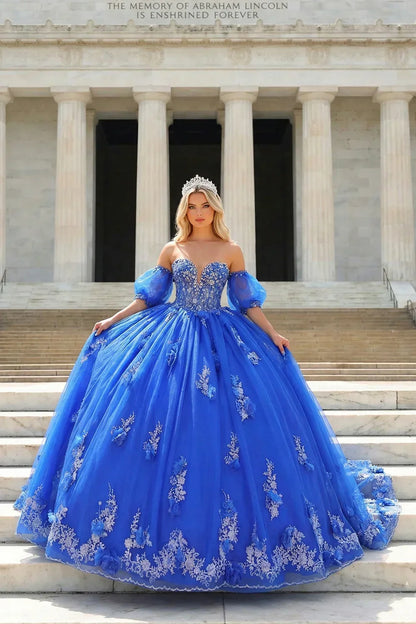 Woman in royal blue off-shoulder ball gown with silver floral embroidery and tiara, standing on marble steps.