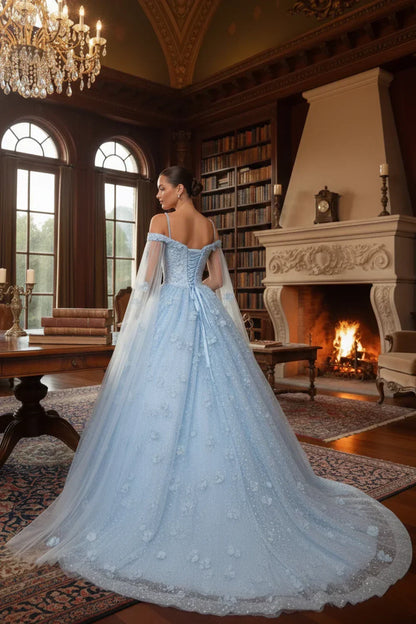Woman in a light blue off-the-shoulder ball gown with sheer cape and 3D floral details in an elegant library setting.