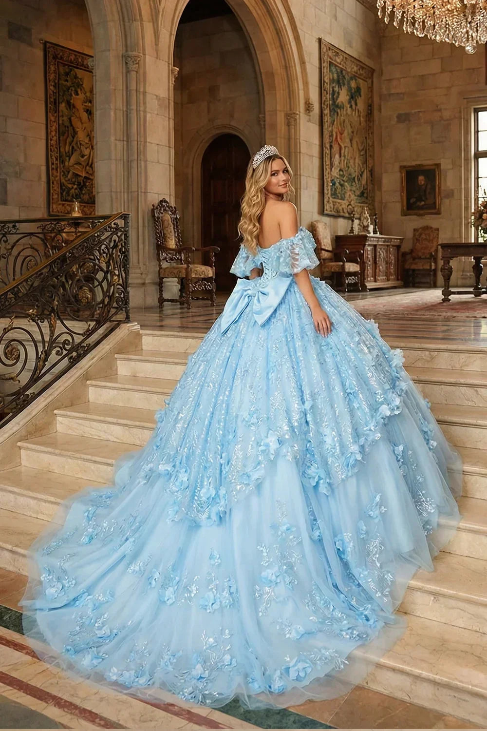 Woman in a light blue ruffle ball gown with 3D floral details and bow on castle staircase
