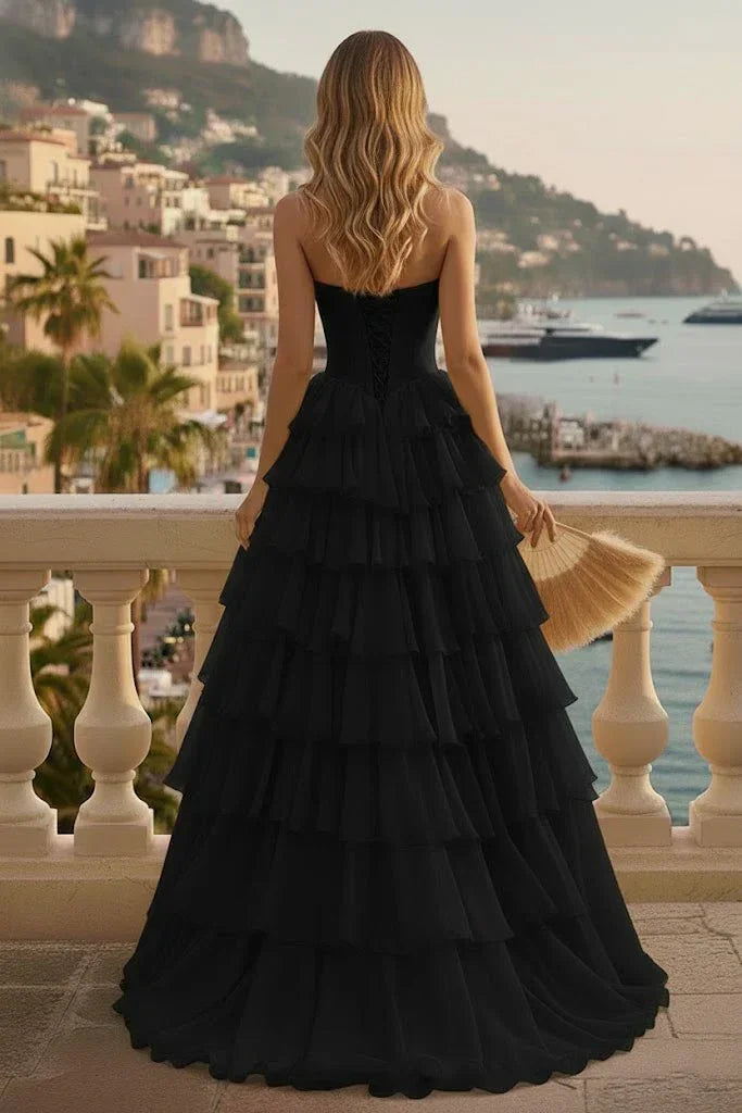 Woman in black tiered ruffle prom dress with lace-up back, standing on a balcony overlooking coastal town
