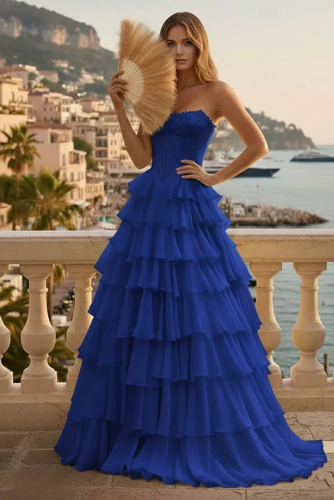 Woman in vibrant blue tiered ruffle corset A-line prom dress with lace-up back on a balcony overlooking a coastal town.