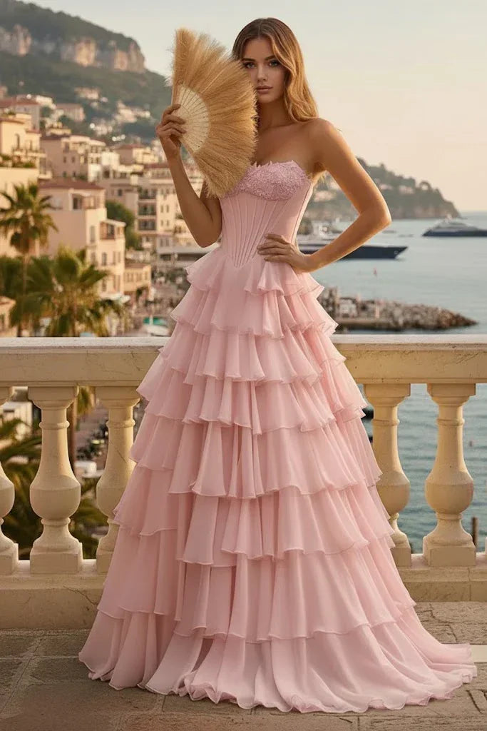 Woman in pink tiered ruffle corset A-line prom dress with lace-up back, standing on a balcony with coastal views.