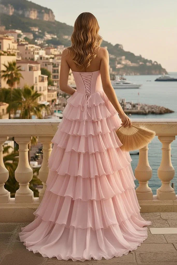 Woman in pink tiered ruffle prom dress with lace-up back, standing on a balcony overlooking the sea